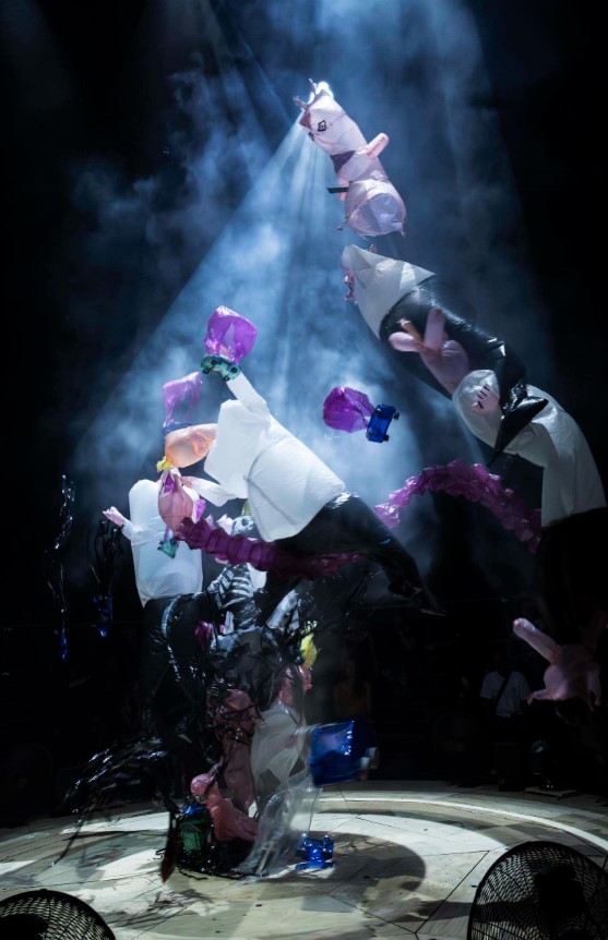 Anthropomorphic plastic and inflatable figures stand beneath a beam of light and smoke.