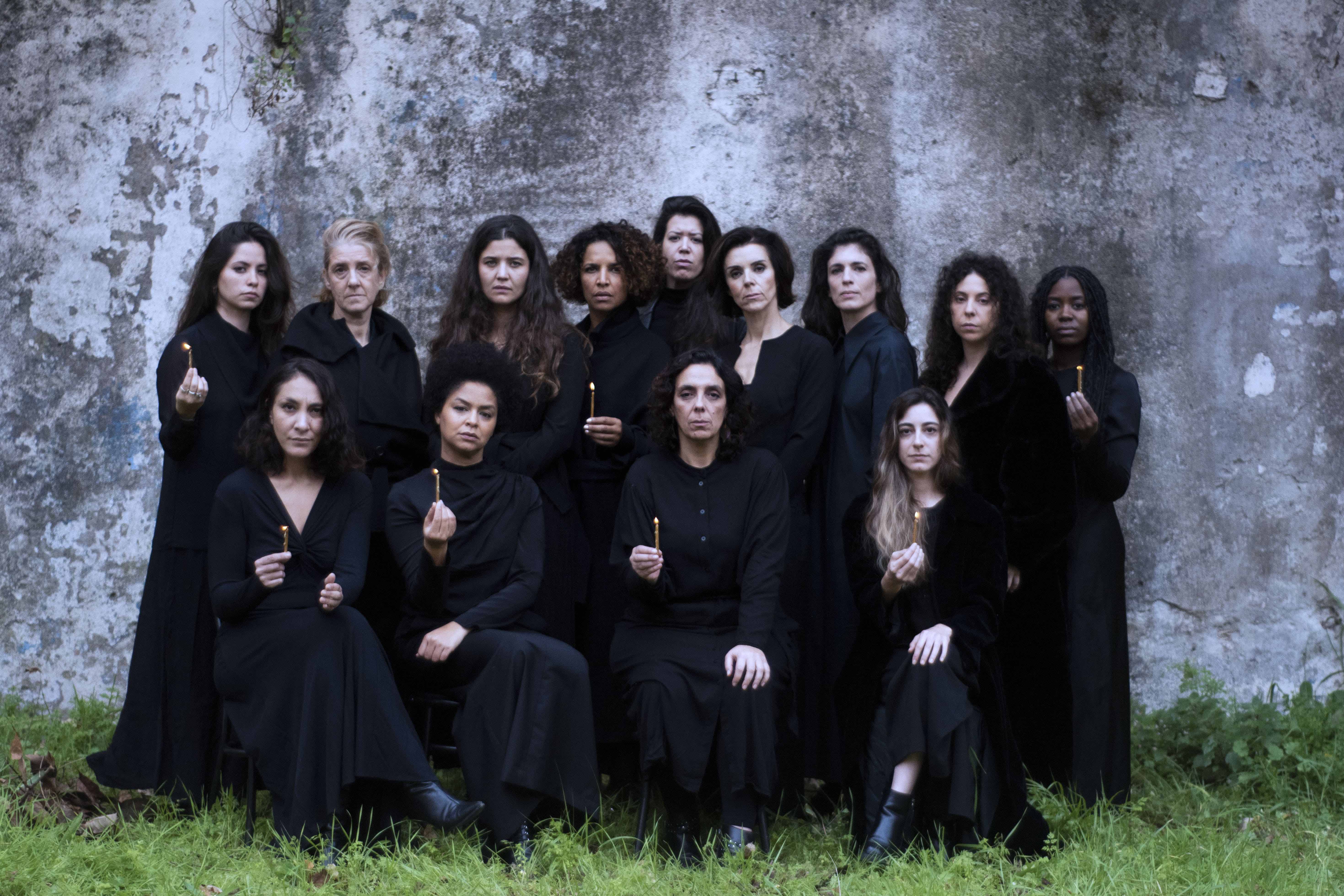 Color photograph of 13 women, some sitting and some standing, all dressed in black, looking towards the camera. Some are holding small candles.