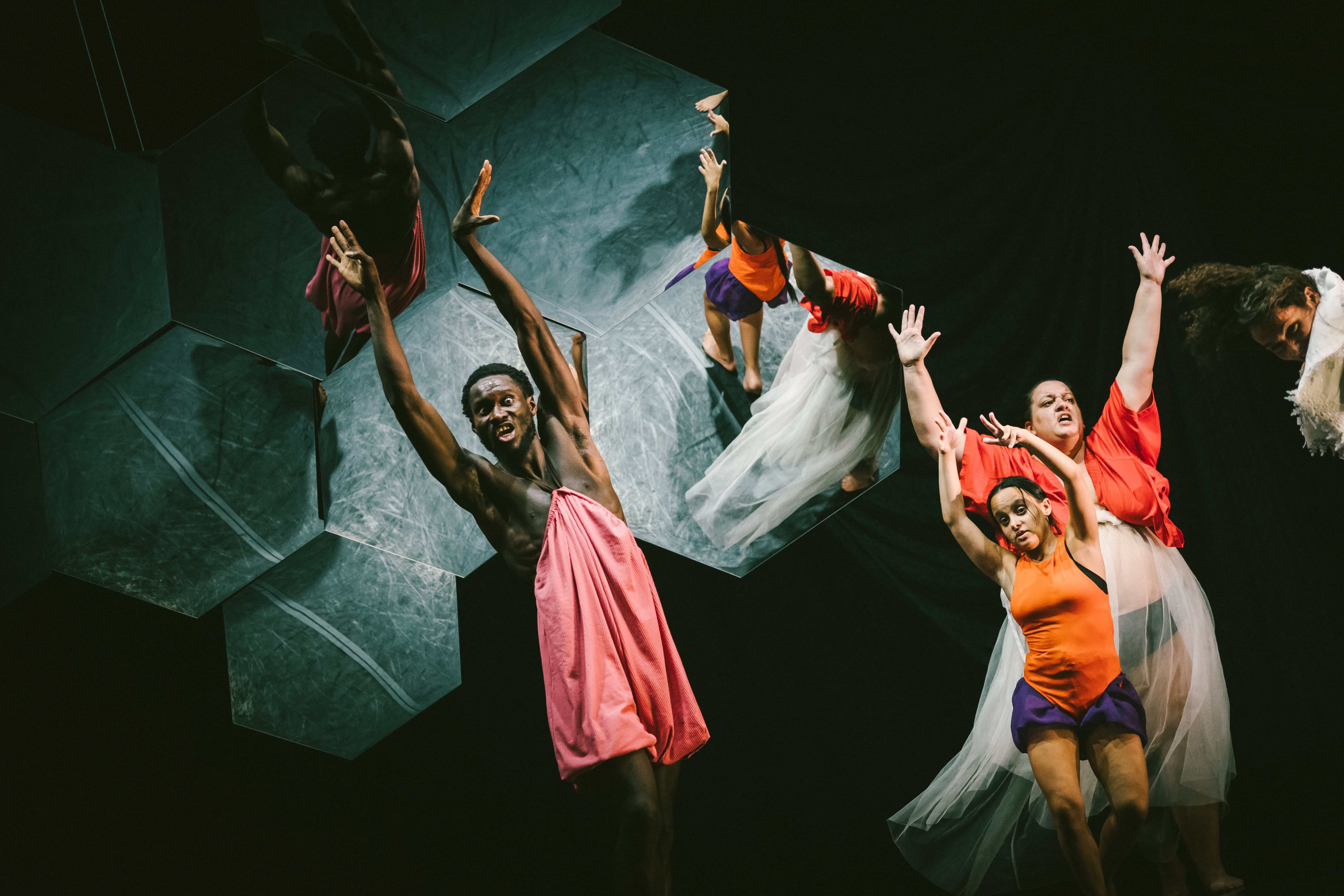Colour stage photo. In the foreground, three performers in colorful clothes raise their arms. In the background, the mosaic of hexagonal mirrors.