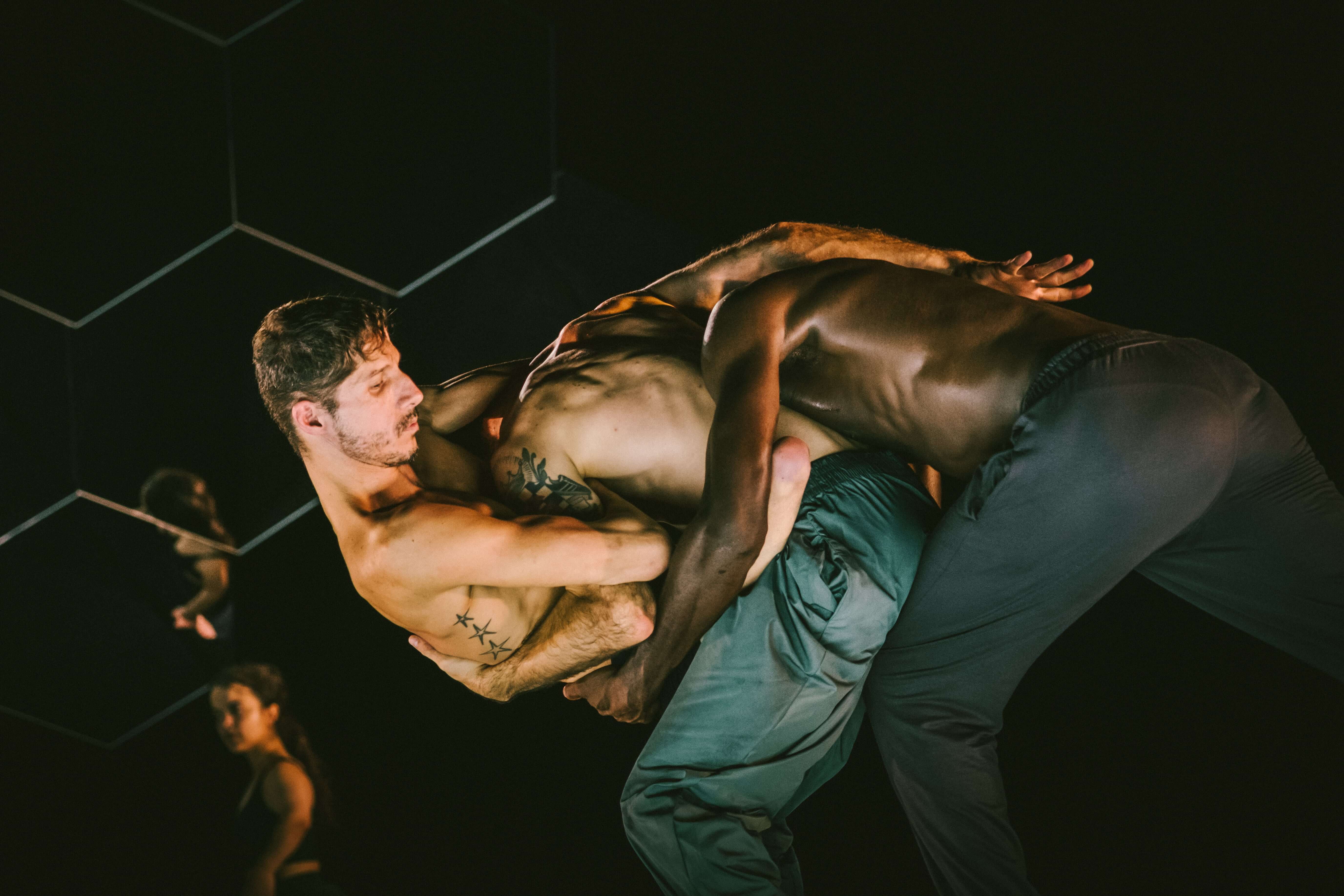 Colour stage photo. Two interpreters cling to each other, back to back,  both backs facing the audience, holding a third performer with their arms.
