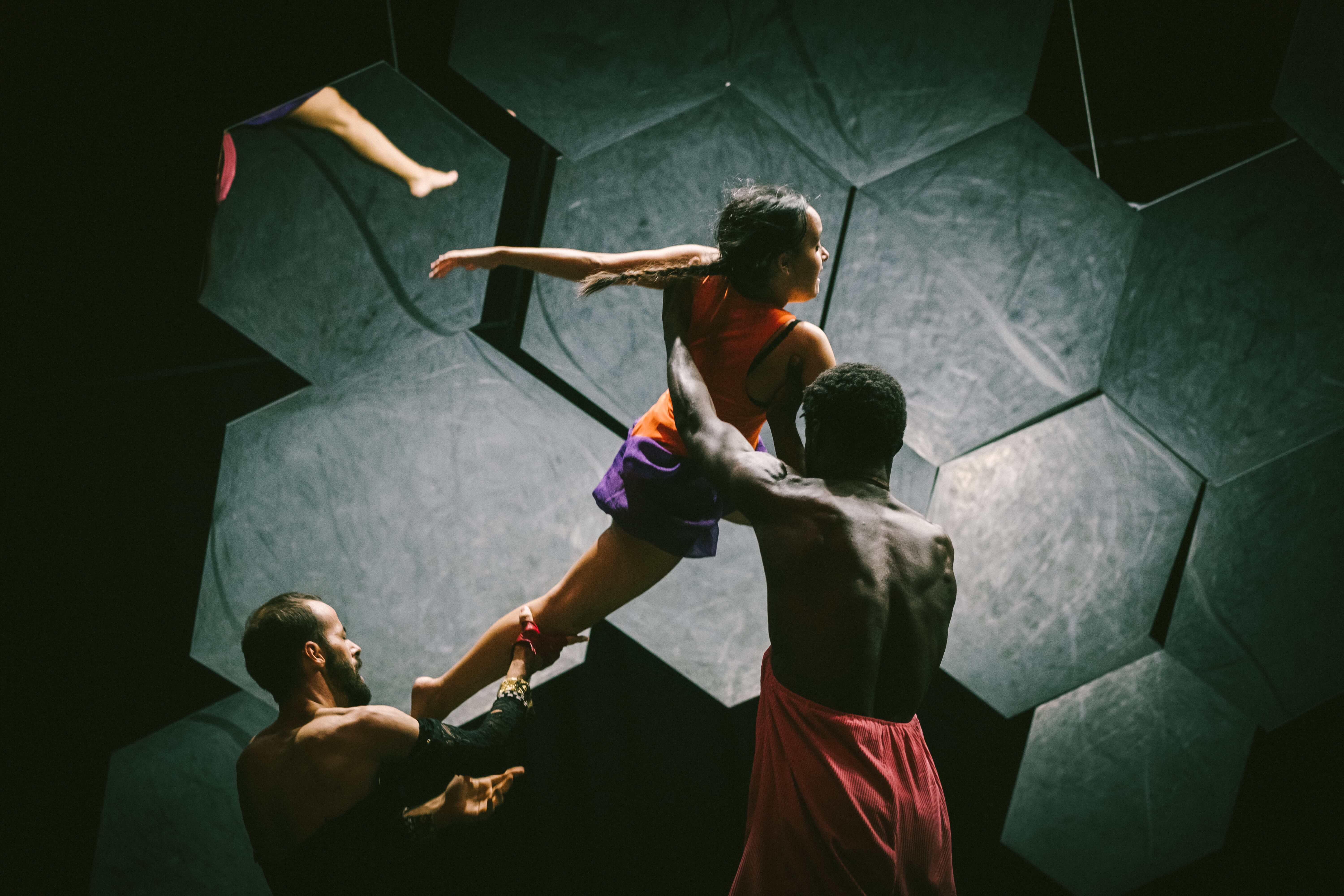 Colour stage photo. Two interpreters lifting a third performer with their arms. In the background, the mosaic of hexagonal mirrors.
