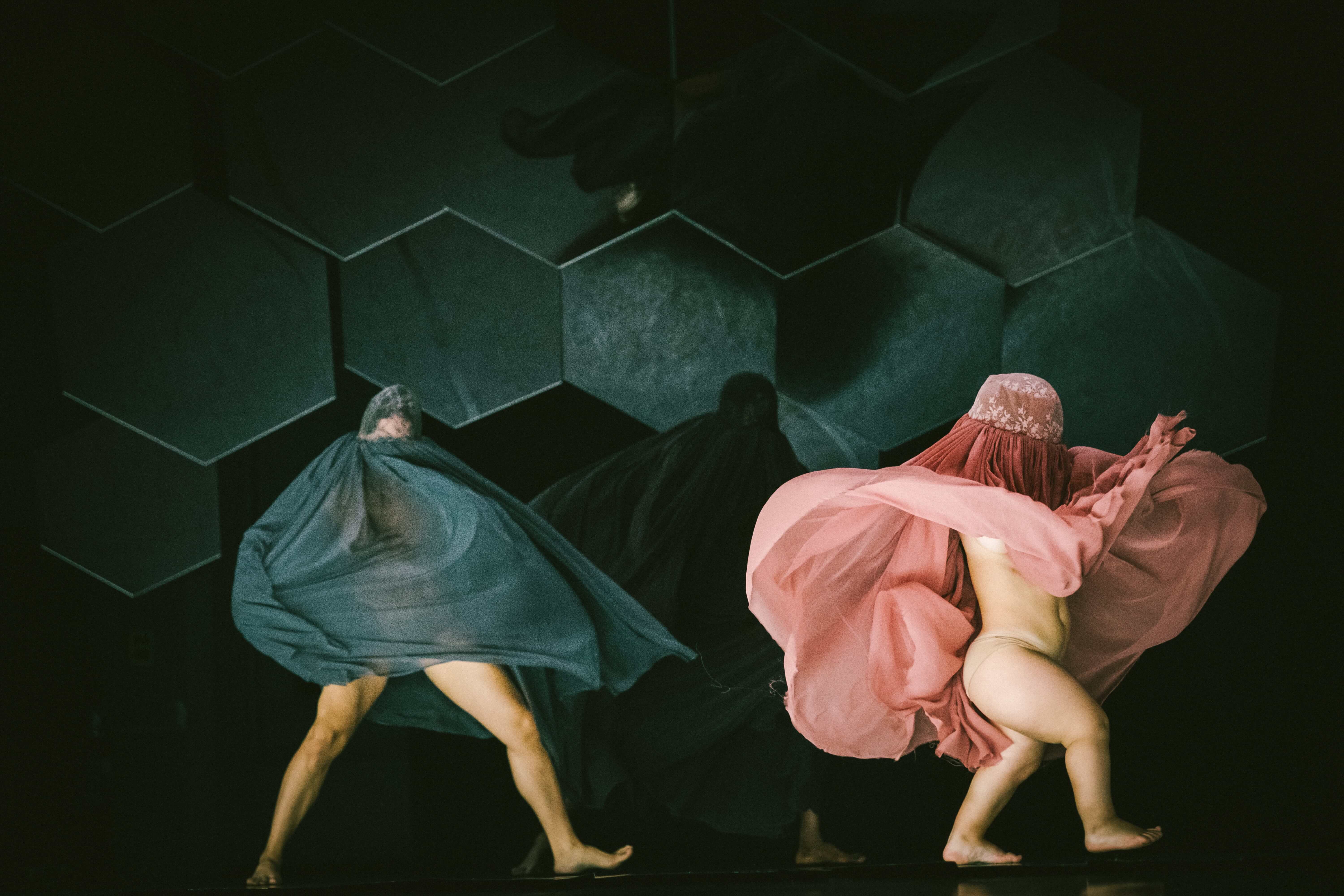 Colour stage photo. In the foreground, two partially covered performers, one with a teal fabric and the other with a light pink fabric. In the gloom, a body fully covered with dark fabric.  